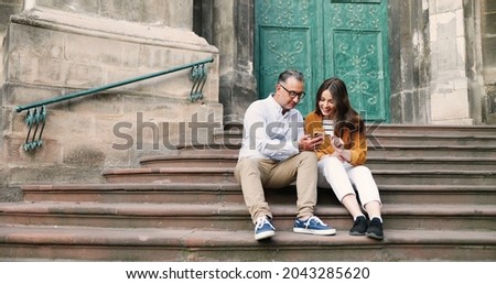 Pretty young Caucasian girl with father sitting on steps of church and using smartphone outdoor. Middle-aged man showing something on mobile phone to beautiful adult daughter, discussing and talking.