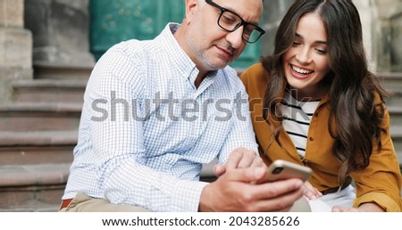 Pretty young Caucasian girl with father sitting on steps of church and using smartphone outdoor. Middle-aged man showing something on mobile phone to beautiful adult daughter, smiling and talking.