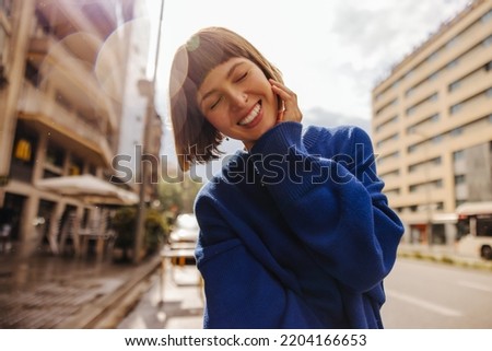 Pretty young caucasian woman closing her eyes smiles teeth walking around city. Brown-haired with bob haircut wears blue sweatshirt. Positive emotions concept Stock photo © 