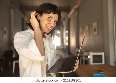 Pretty Young Caucasian Woman Looks Into Camera, Tucking Hair Behind Ear Holding Laptop. Brunette Wears White Shirt Standing Indoors. Concept Of Technological Device.