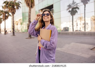 Pretty young caucasian woman with notebooks walks around city after class. Brown-haired girl wears sunglasses and purple shirt. Student concept