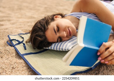 Pretty young caucasian woman reading book relaxing on beach in sunny weather. Model wears white-blue sundress combines business with pleasure. Relaxing mood concept. - Shutterstock ID 2254209851