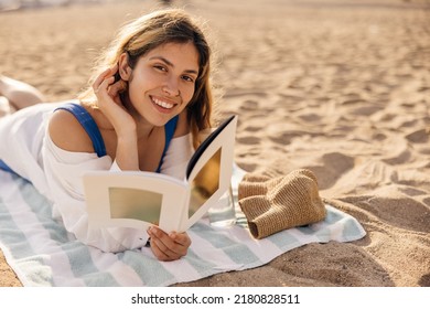 Pretty young caucasian woman spends leisure time reading book lying on beach sunny day. Brown-haired girl smiling looks at camera, wears shirt. Weekend enjoyment concept - Powered by Shutterstock