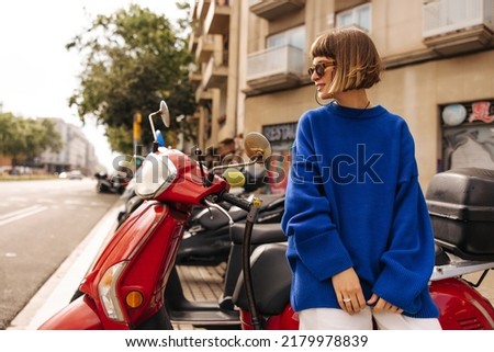 Pretty young caucasian woman in sunglasses sits on moped and looks away at view of city. Brown-haired with a bob haircut wears blue sweater in autumn. City life concept Stock photo © 