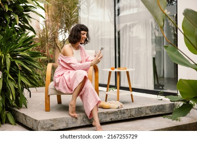 Pretty Young Caucasian Woman Is Talking On Video Link Through Phone Sitting In Patio. Brunette With Short Haircut Wears Pink Summer Suit. Positive Emotions Concept