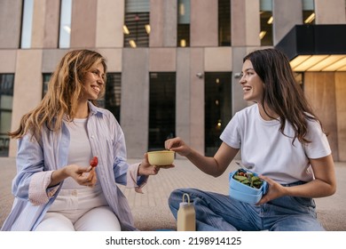 Pretty young caucasian women eat bowl salad sitting on stairs in street near office building. Blonde with brunette wear casual clothes. Concept of enjoying moment