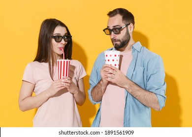 Pretty Young Couple Two Friends Man Woman In 3d Glasses Isolated On Yellow Background. People Emotions In Cinema Lifestyle Concept. Watching Movie Film Hold Cups Of Soda Or Cola, Looking At Each Other