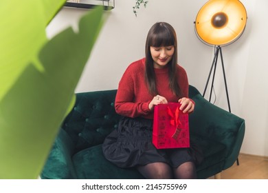 Pretty Young Dark Haired Woman Opening A Present In Red Bad Birthday Home Background Medium Full Shot Copy Space. High Quality Photo