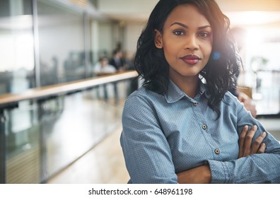 Pretty young dreamy African-American office worker standing with arms crossed and looking at camera.