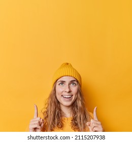 Pretty young European woman points fingers up smiles happily shows advertisement demonstrates logo or company banner upwards wears hat and casual comfortable jumper isolated over yellow background