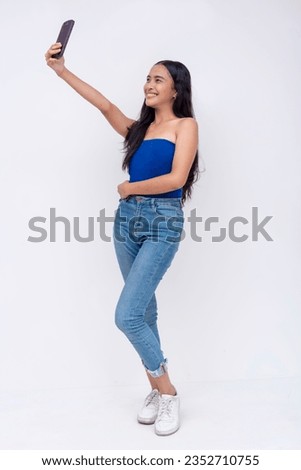 A pretty young expressive asian woman takes an upward angled selfie of herself with her phone. A modern trendy gen z lady. Full body photo isolated on a white backdrop.