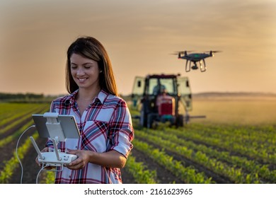 Pretty young farmer woman driving drone with remote control in front of tractor in soybean field in spring at evening - Powered by Shutterstock