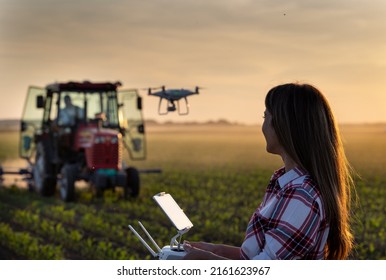 Pretty young farmer woman driving drone with remote control in front of tractor in corn field in spring at evening - Powered by Shutterstock