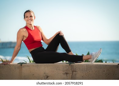 Pretty Young Fit Athletic Blonde Woman In Yoga Pants And Red Shirt Sitting Posing On Beach At Sunset With Blonde Hair