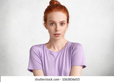 Pretty young ginger female with hair knot, dressed in light casual purple t shirt, looks confidently at camera, isolated over white concrete wall. Cute red haired woman rests alone after work