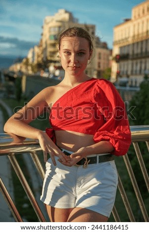 pretty young girl with afternoon sunlight bathing her face, dressed in a red one-shoulder shirt and white shorts, posing on the railing of a bridge with the city in the background.
