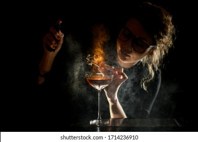 Pretty young girl bartender at dark bar carefully decorates white flowering branch with glass of drink using tweezers and sprinkles on it
