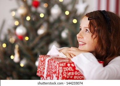 Pretty young girl dreaming of Christmas as she rests her chin on top of a decorative gift in colorful red wrapping with a sparkling Christmas tree behind