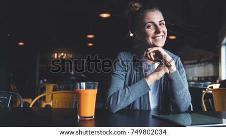 Pretty young girl with happy smile using mobile tablet during taking lunch brake, drinking bright fresh orange juice. Healthy food in cafe. Holding glasses.