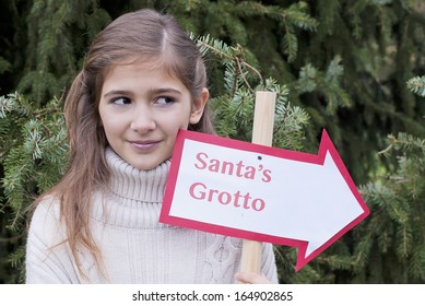 Pretty, Young Girl Holding A Santa's Grotto Sign In Front Of A Fir Tree