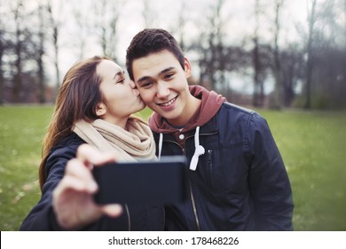 Pretty young girl kissing her boyfriend on cheeks while taking self portrait with a mobile phone. Mixed race couple in park.