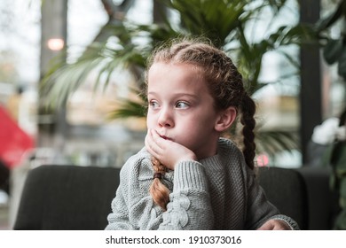 Pretty young girl with pigtails and grey wooly jumper sat in modern school environment looking out of window to natural light day dreaming with head resting on childs hand palm plant tree behind