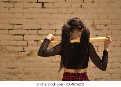 Pretty young girl with pigtails and punk style with a baseball bat resting behind her neck on her back on a yellow brick wall in the background.