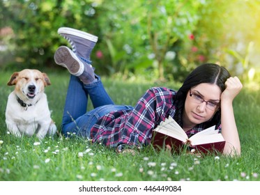 Pretty young girl reading book on grass in park with dog beside her