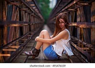 Pretty And Young Girl Sitting On A Rusty Metal Bridge. She Wears A Blue Jeans Shirts And White T-shirt. The Wind Blowing In Her Long Hair. A Fashion Photography.