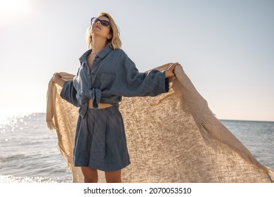  Pretty young girl stands on seashore with her head tilted back, developing blanket behind her back. Blonde in relaxed state is resting, basking in sun in blue skirt and shirt tied at her belly.