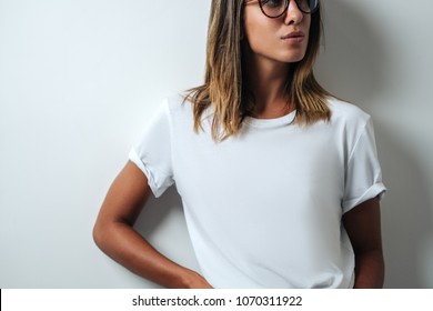 Pretty young girl in white blank t-shirt, wearing glasses, empty wall, studio portrait