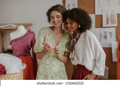 Pretty young girls look at phone screen. Ladies in stylish summer outfits smile and pose in cozy office of fashion designer. - Powered by Shutterstock