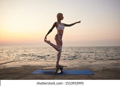 Pretty Young Healthy Woman With Casual Hairstyle Holding Pose On Balance Board On Seafront, Posing In Sporty Clothes Over Sea View During Sunrise, Keeping Balance On Wooden Desk