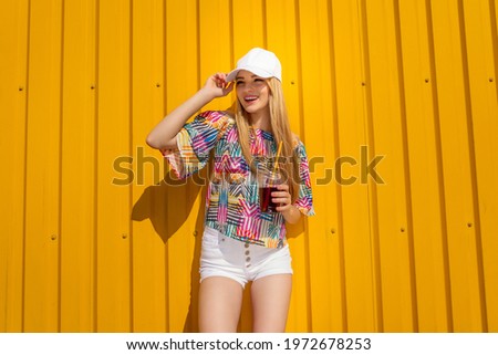 Pretty young hipster style girl have fun outdoor in white cap and eating ice cream on yellow background urban style. 
