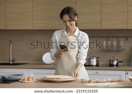 Pretty young housewife in apron holds cellphone, check on-line cookbook recipe, makes photo of fresh baked prepared pizza for new blog post in social media network enjoy cooking time and hobby at home