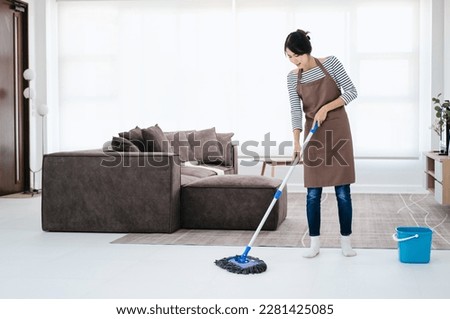 Pretty young lady in apron wiping floor with mop, doing house cleanup living room, free space. Positive housewife tidying her apartment, doing household chores. Professional sanitary service concept.
