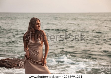 Pretty young lady in beige dress relaxing on beach at tropical ocean background, looking away. Cute slender woman enjoy rest on seacoast. Travel vacation holiday concept. Copy advertising text space