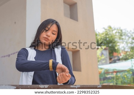 A pretty young lady wearing a cardigan and a navy blue long sleeves top is checking the time on her gold colored wrist watch. Plants shown in the background
