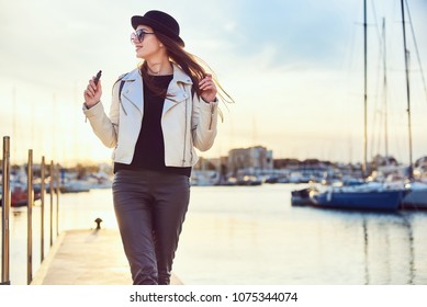 Pretty young long-haired woman blogger in black hat, round sunglasses holding vape, smiling and walking at the sunset in a port with sea and boats on background.