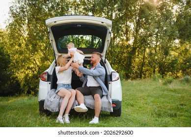 Pretty young married couple and their daughter are resting in the nature. The woman and girl are sitting on open car boot.