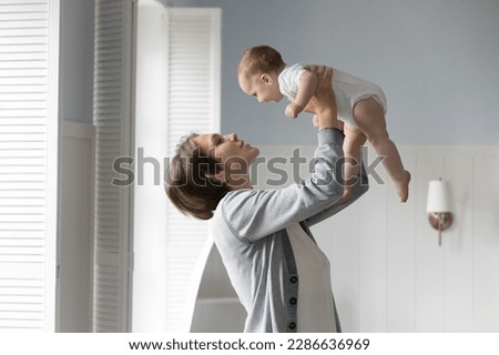 Pretty young mom lifting infant baby up in air, throwing, holding little child in arms, playing with cute kid with love, tenderness, devotion, enjoying new motherhood at home