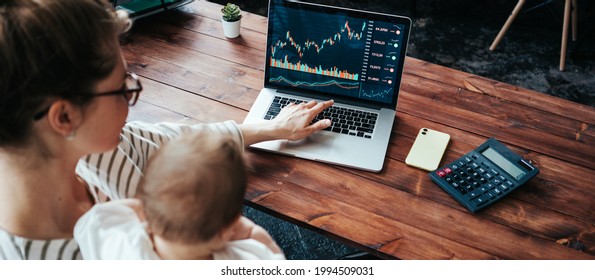 Pretty Young Mother With Cute Little Baby Investing In World Stock Market, Using Her Laptop And Online Trading Soft From Home