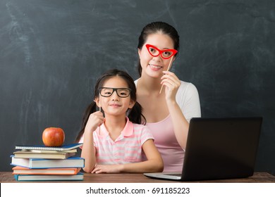 Pretty Young Mother With Cute Little Daughter Studying With Computer Together Prepare Back To School And Using Glasses Toy Dress Up Looking At Camera In Blackboard Background.