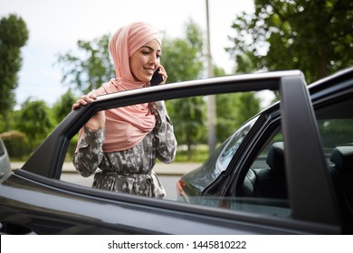 Pretty Young Muslim Woman In Traditional Dress Talking On Phone When Getting Into Car