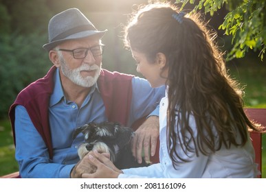 Pretty Young Nurse In White Coat Holding Senior Man For Hand In Park. Cute Dog Sitting In Lap On Bench