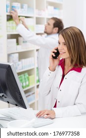 Pretty Young Pharmacist Chatting On The Phone As She Checks Information On The Computer While A Male Colleague Works In The Background