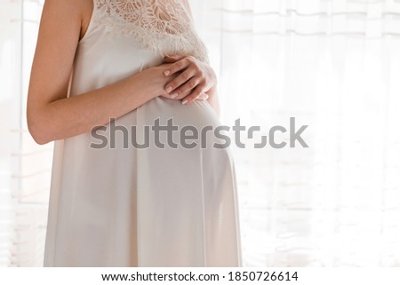 Pretty young pregnant woman standing by the window in home.