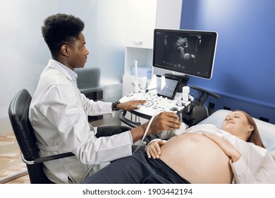Pretty young pregnant woman visiting her doctor for ultrasound examination. Professional male African doctor sonographer performing 3d ultrasound on pregnant woman in modern clinic