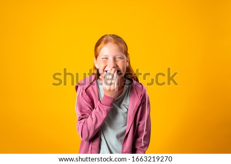 A pretty young red haired teenager covers her mouth with her hand while nervously laughing. Cute preteen Caucasian girl giggles with her hand close to her lips. Concept of fun, pranks, joking around Foto d'archivio © 