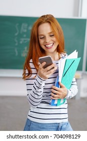 Pretty Young Redhead Student Reading An Sms Or Text Message On Her Mobile Phone With A Smile As She Carries Folders In The Classroom